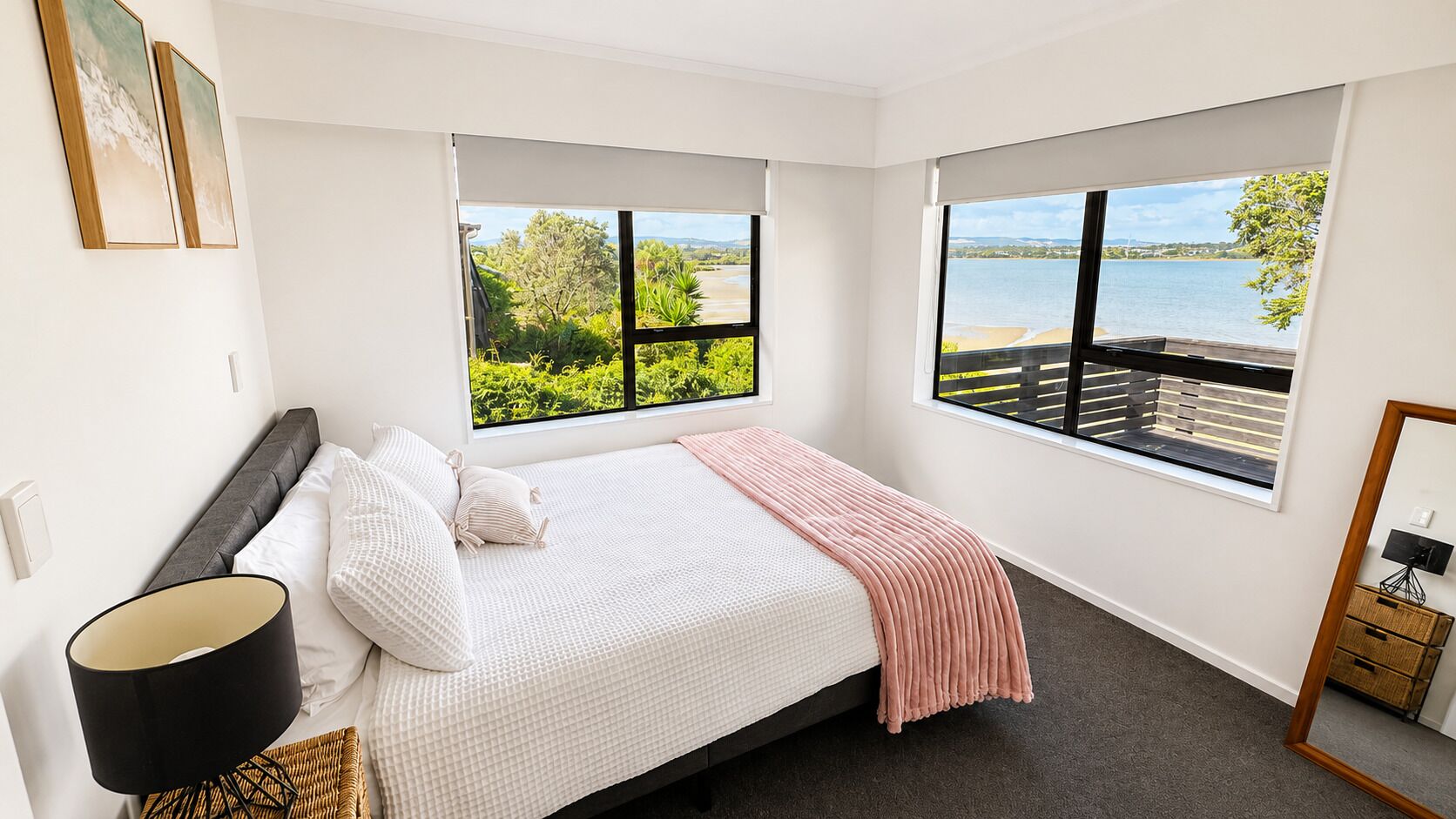 A bedroom with a bed, a nightstand, a lamp, and a view of a body of water through large windows.