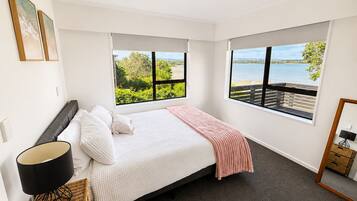 A bedroom with a bed, a nightstand, a lamp, and a view of a body of water through large windows.