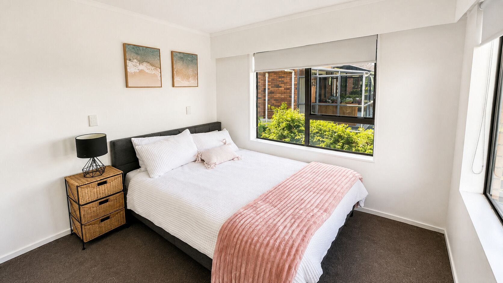 A bedroom with a bed, a bedside table with a lamp, a window with a view of greenery, and two wall-mounted pictures.