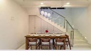 A dining area with a wooden table set for four, a staircase with glass railings, and a light blue wall. - Tranq Villa by Stay Serene Luxury (Murbad)
