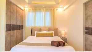 A hotel room with a large bed, a nightstand with a lamp, and a window with curtains. - Tranq Villa by Stay Serene Luxury (Murbad)