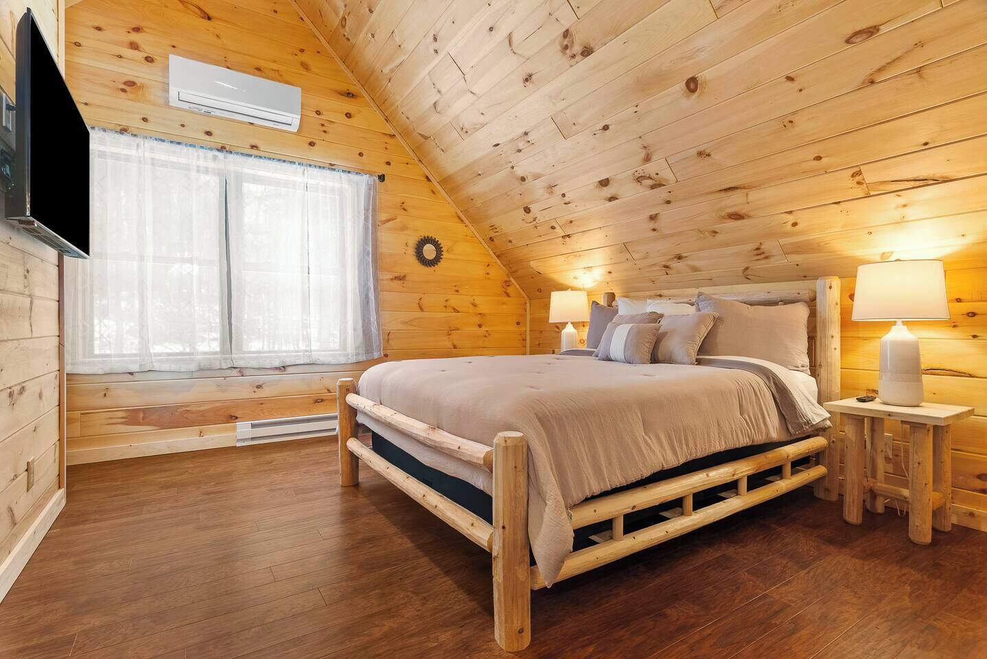 Un dormitorio acogedor tipo cabaña con cama, dos lámparas de noche, una ventana con cortinas traslúcidas y un aire acondicionado instalado en la pared.