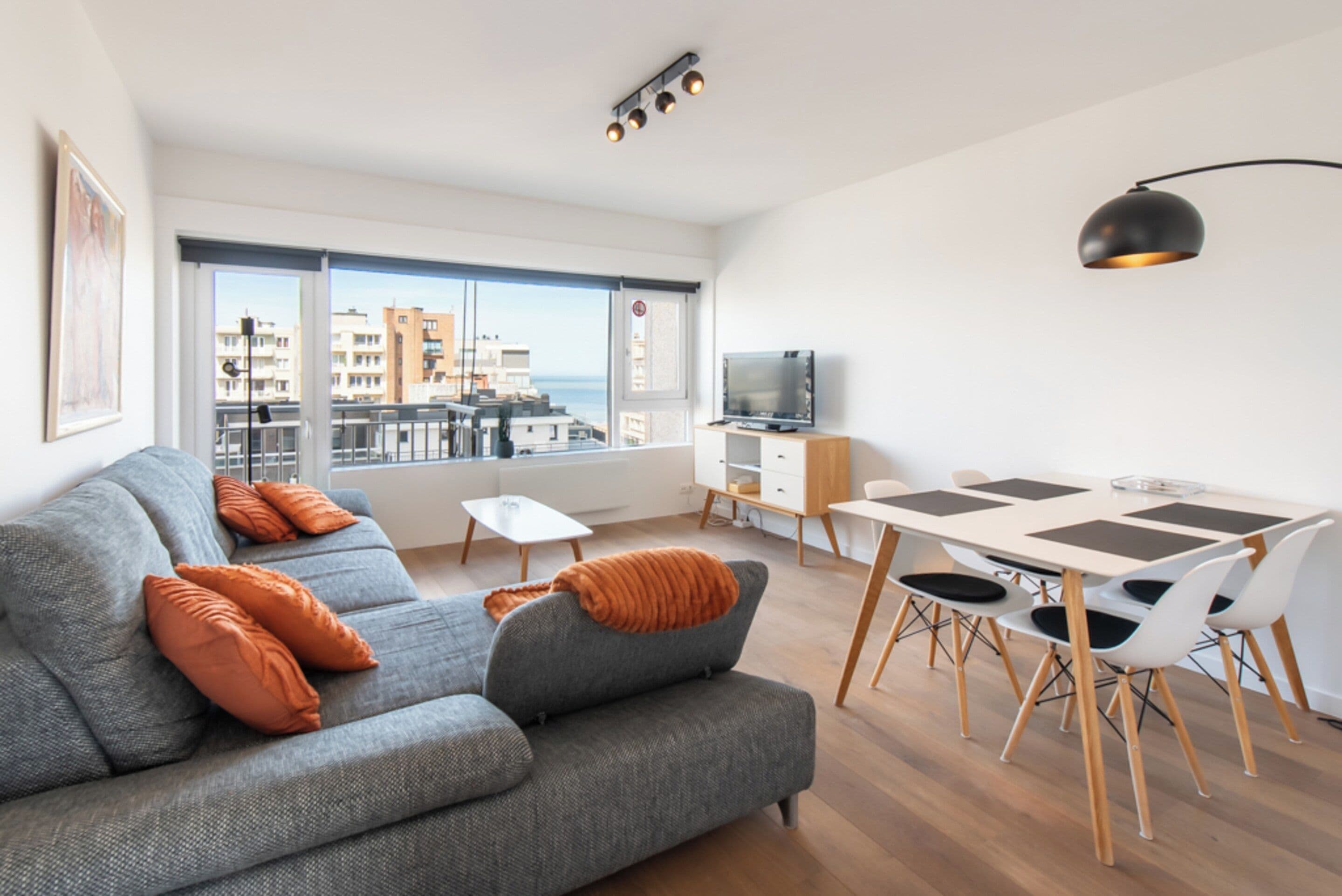 A modern living room with a grey sofa, orange cushions, a dining table with chairs, a TV on a stand, and a view of buildings through large windows.