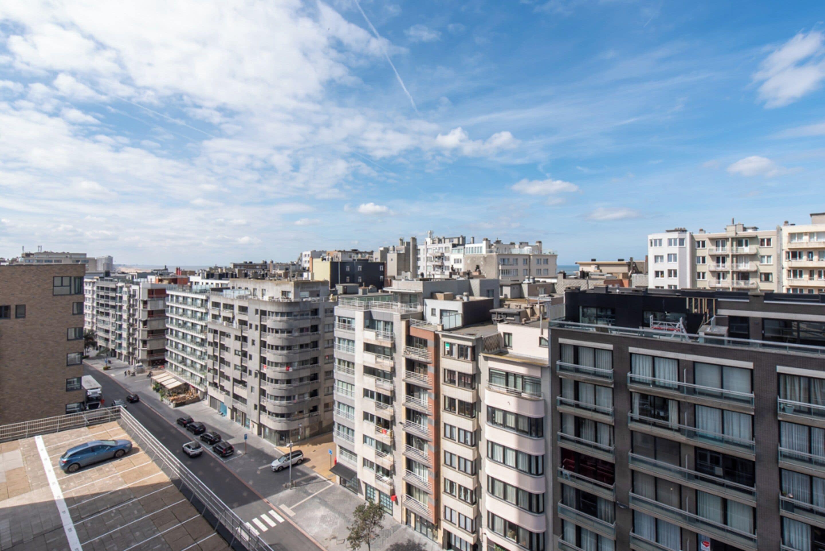 A high-rise residential building with multiple balconies, a parking area, and a clear sky.
