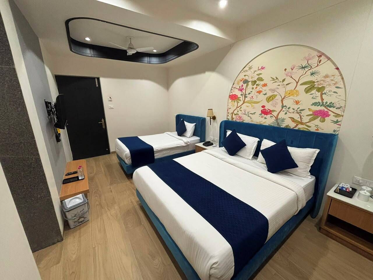 Habitación de hotel con dos camas, un ventilador de techo, un reloj en la pared y una mesita de noche pequeña con una lámpara.