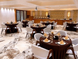 Un comedor de hotel con mesas redondas, sillas blancas y una barra de bar al fondo.