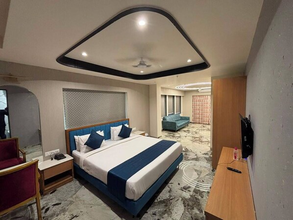 A hotel room with a bed, a desk, a chair, a TV, and a ceiling fan. - Hotel Royal Galleria (Vadodara)