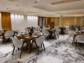 A dining area with multiple tables and chairs, a kitchen door, and patterned wallpaper. - Hotel Royal Galleria (Vadodara)