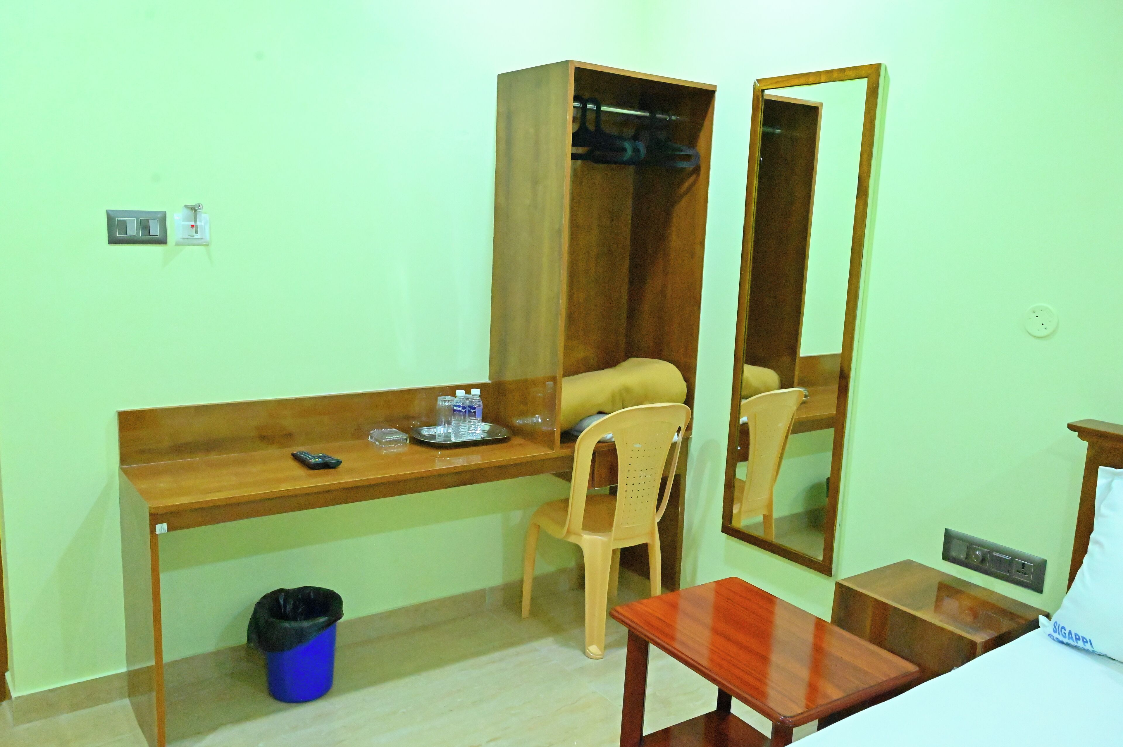Una habitación pequeña con un escritorio de madera, una silla, un espejo, un cubo de basura y una cama.