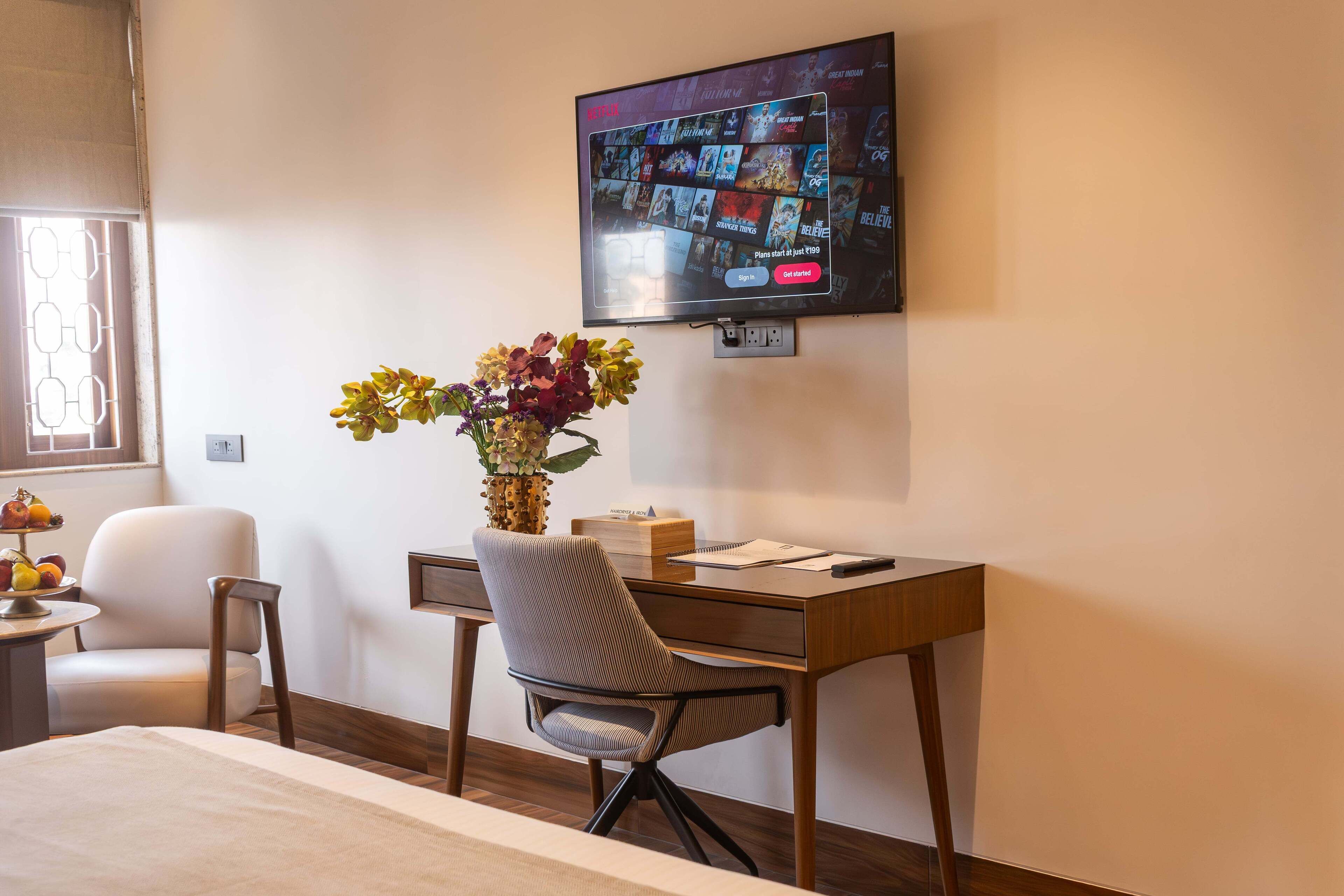 A hotel room with a bed, a desk with a chair, a television mounted on the wall, and a vase with flowers on the desk.