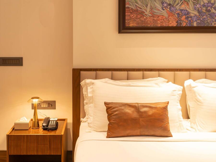 A neatly made bed with white linens and a single brown pillow, a bedside lamp, a telephone, and a small box on a wooden nightstand.