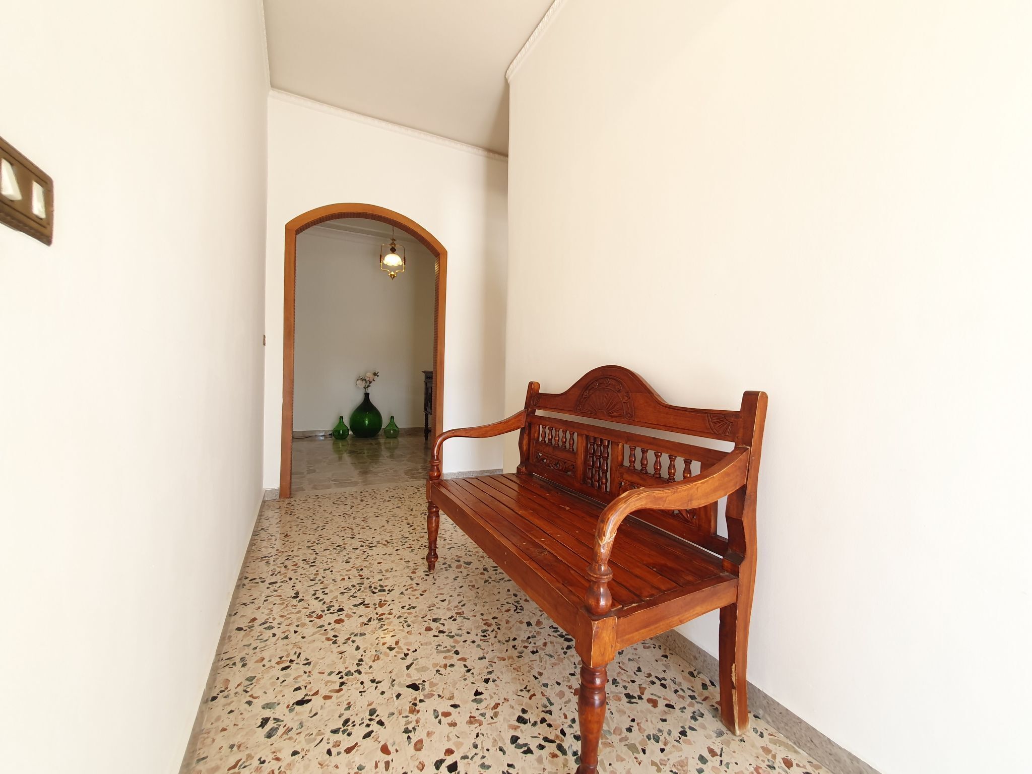 Un banc en bois dans un couloir, avec un sol à motifs et un vase vert rempli de fleurs en arrière-plan.