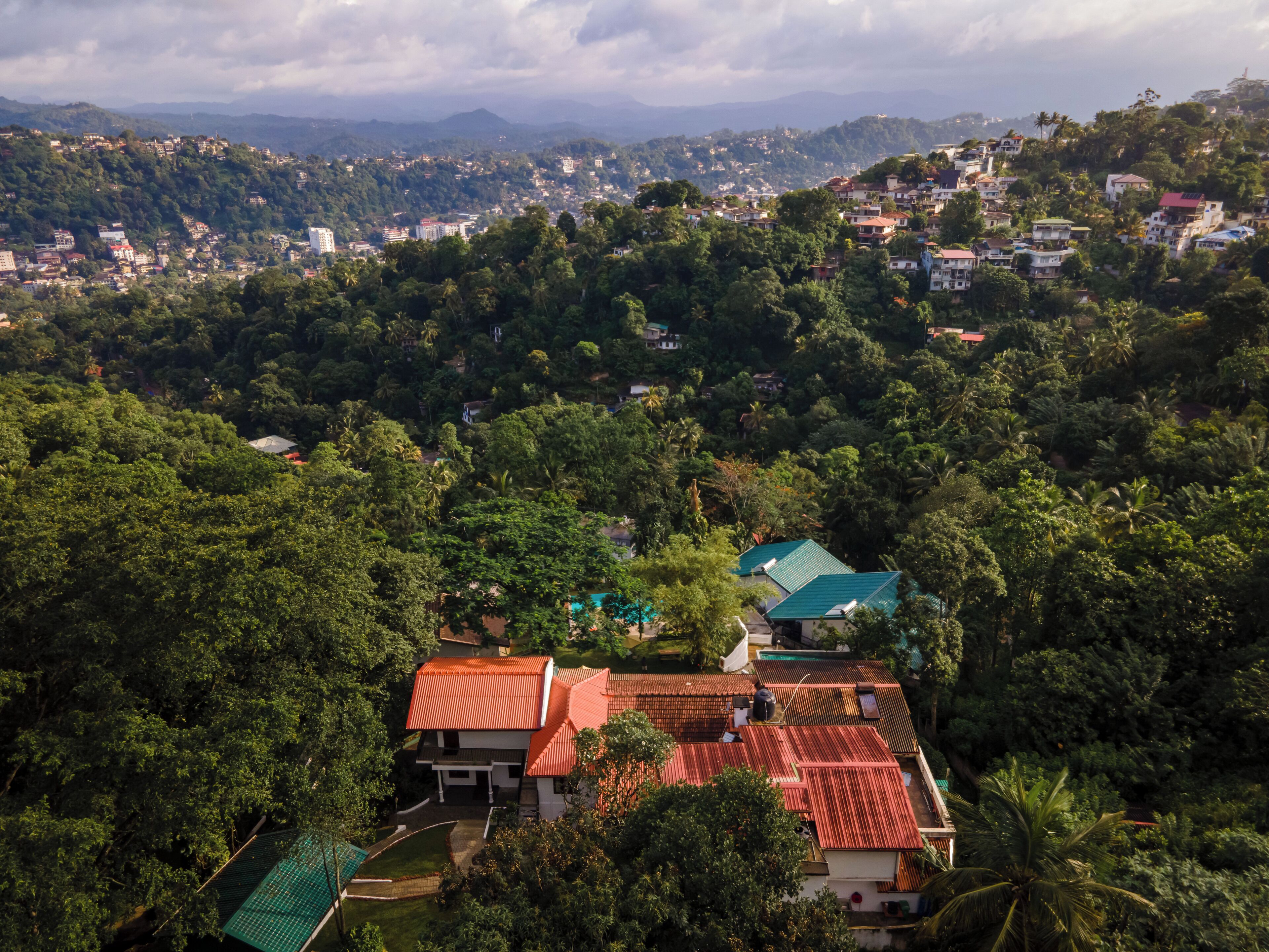 A residential area with houses surrounded by dense greenery and hills in the background.