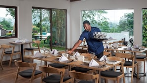 A restaurant with wooden tables and chairs, large windows with a view of greenery, and a waiter serving a tray. - Mirage Kandyan Heights (Kandy)