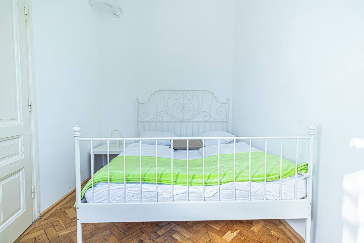 Una cama de metal blanco con una manta verde en una habitación con paredes blancas y suelo de madera.