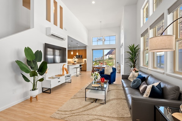 A spacious living area with a modern sofa, a coffee table, and a large plant. There is a kitchen in the background with a dining area.