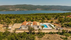 En moderne villa med swimmingpool, omgivet af grøn vegetation og udsigt over et sø med vindmøller i baggrunden.