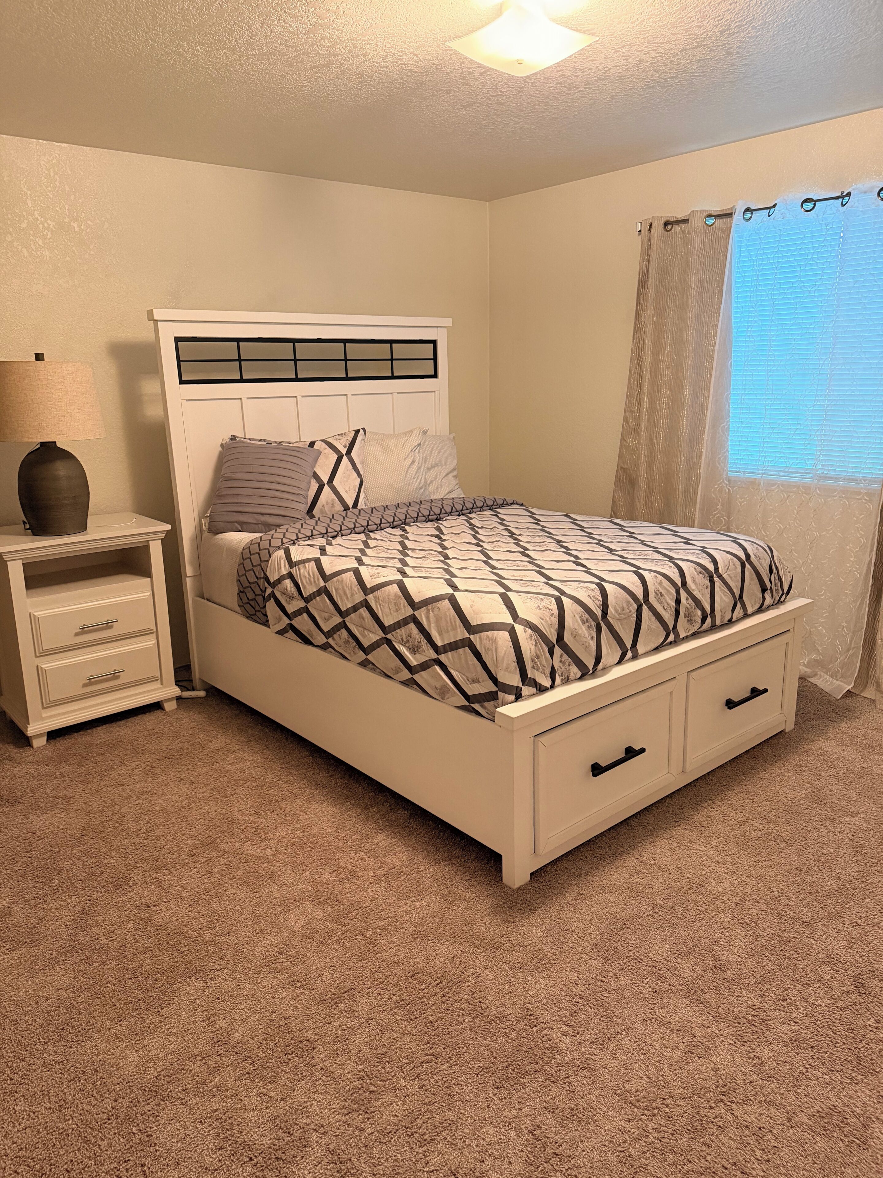 A bedroom with a bed, nightstand, lamp, and window with curtains.