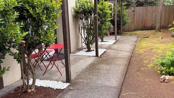 A covered outdoor patio with a concrete walkway, red folding chairs, and small trees.