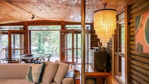 A cozy cabin interior with a wooden ceiling, a dining area with a table and chairs, and a large window offering a view of greenery outside. - Pool, stroll to beach and cafe (PEARL BEACH)
