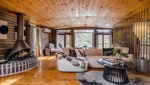 A cozy log cabin interior with a fireplace, wooden ceiling, and large windows offering a view of the outdoors. - Pool, stroll to beach and cafe (PEARL BEACH)