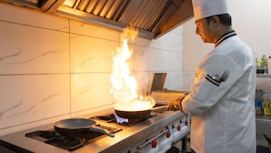 A chef cooking with a pan on a stove, a kitchen counter, and a tiled backsplash. - Aliste millennium City centre, Fortis, Gurugram (Gurugram)