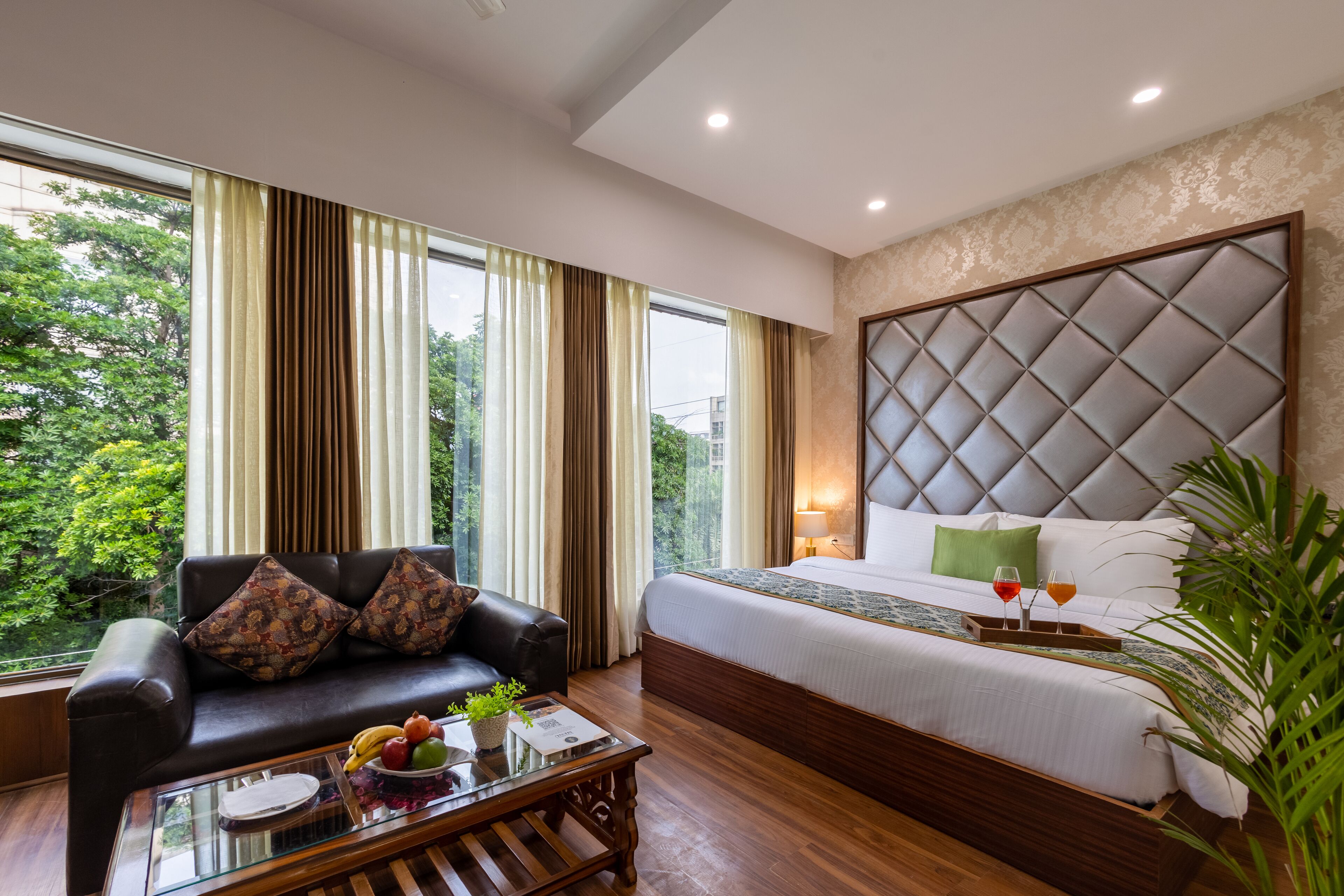 A hotel room with a large bed, a leather sofa, a glass coffee table with fruits, and a view of greenery through the window.