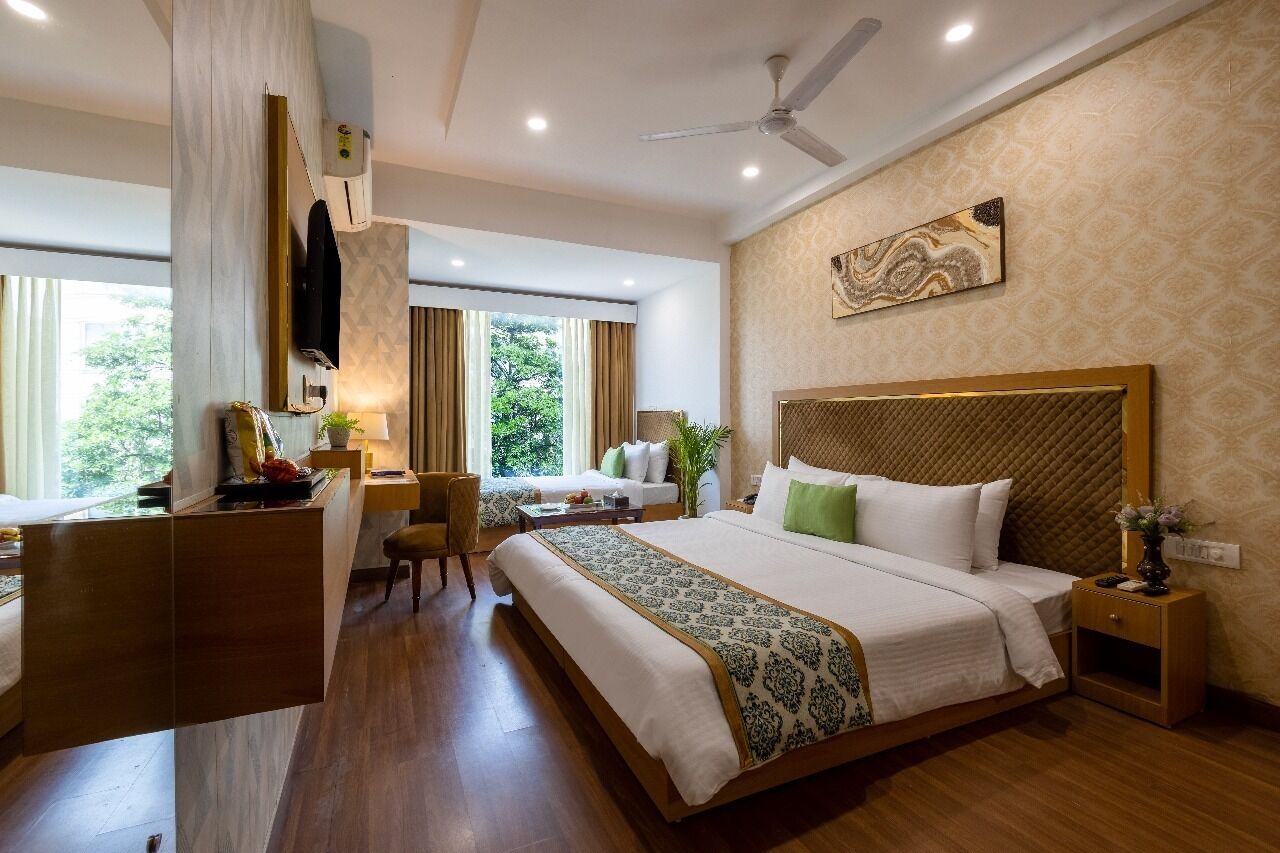 A hotel room with a large bed, a desk, and a chair. There is a window with curtains, a ceiling fan, and a wall-mounted TV.