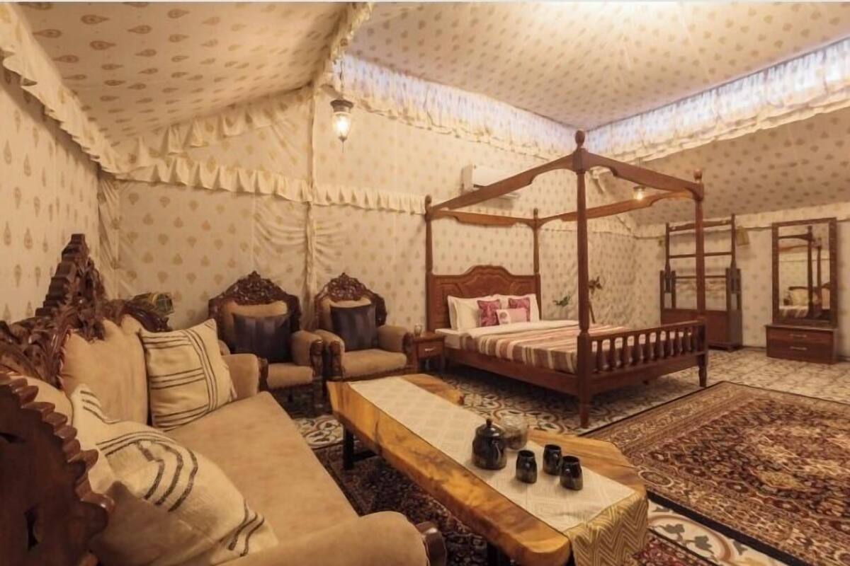 Una cama tradicional con dosel y cuatro postes, un sofá, una mesa de centro de madera y una alfombra estampada en una habitación con papel tapiz con diseño y un ventilador de techo.