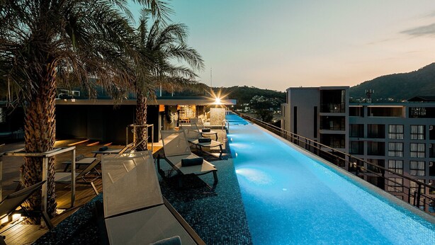 Un espace aménagé au bord de la piscine, avec des chaises longues, un palmier et un bâtiment doté de plusieurs balcons.
