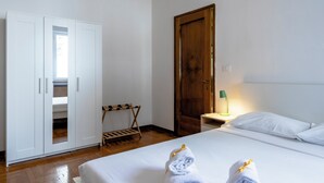 A hotel room with a bed, a wooden wardrobe, a wooden door, a nightstand with a lamp, and a towel rack.