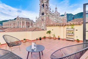 Une terrasse sur le toit, avec des chaises en osier, une petite table et des plantes en pot. Au fond, on aperçoit une grande église à l’architecture riche, dotée d’un clocher.