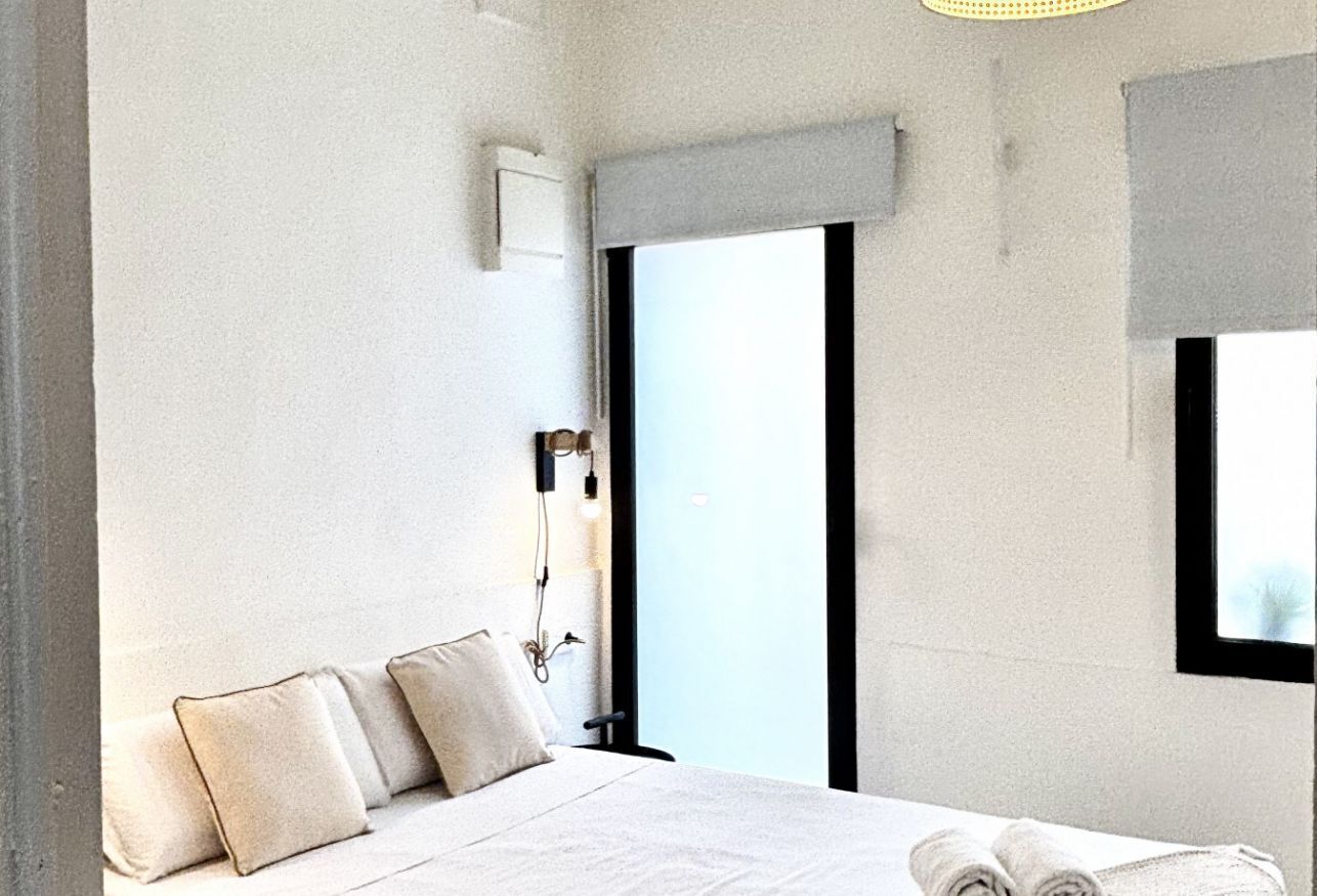 Una habitación de hotel moderna con una cama grande, un ventanal con vistas, una lámpara de pared y una mesita de noche pequeña.