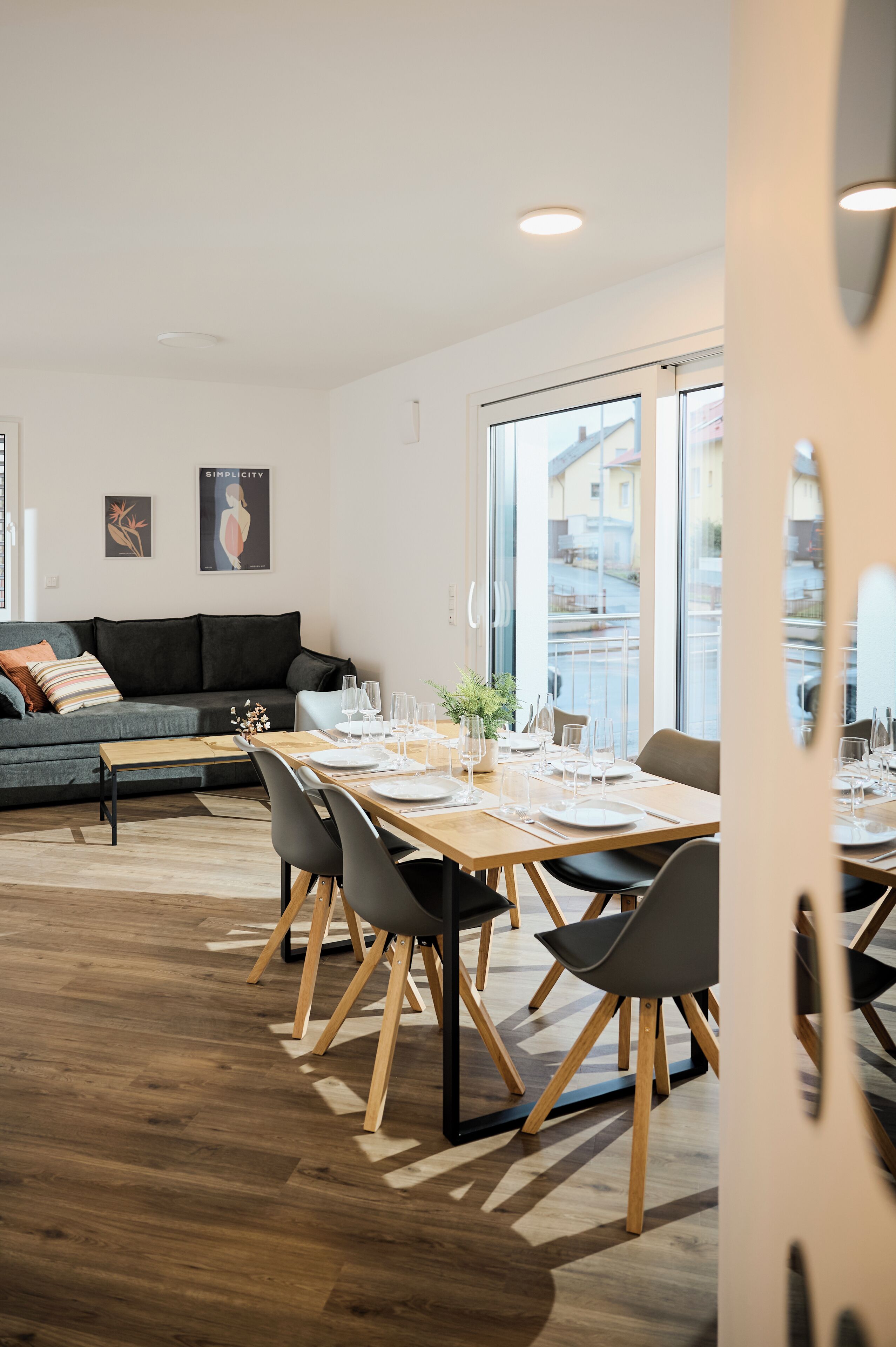 A modern living room with a dining area, featuring a sofa, a wooden dining table set for four, and large windows offering a view of the outside.