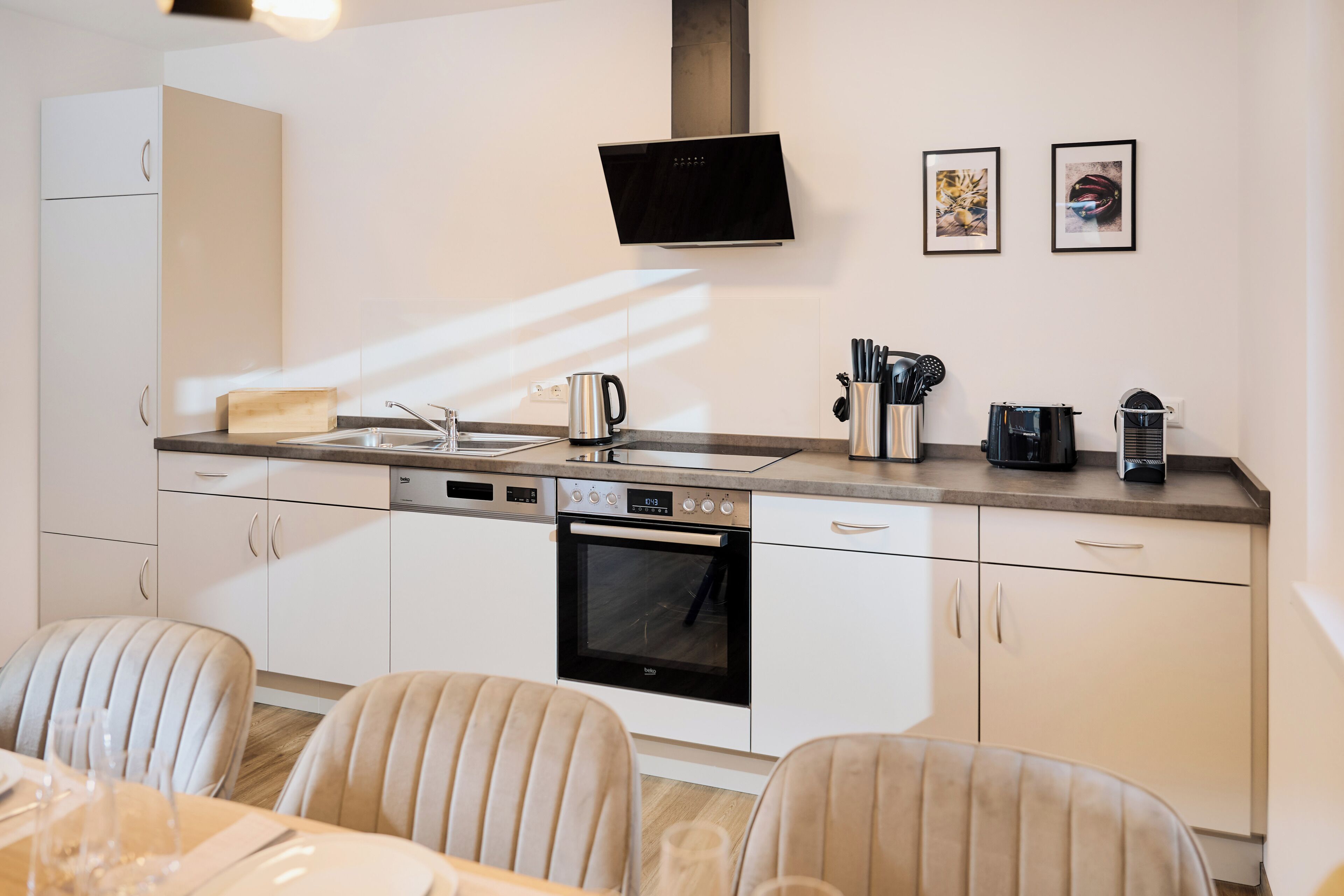 A modern kitchen with white cabinetry, a built-in oven, a dishwasher, and a coffee maker. There are two framed pictures on the wall and a flat-screen TV above the counter.