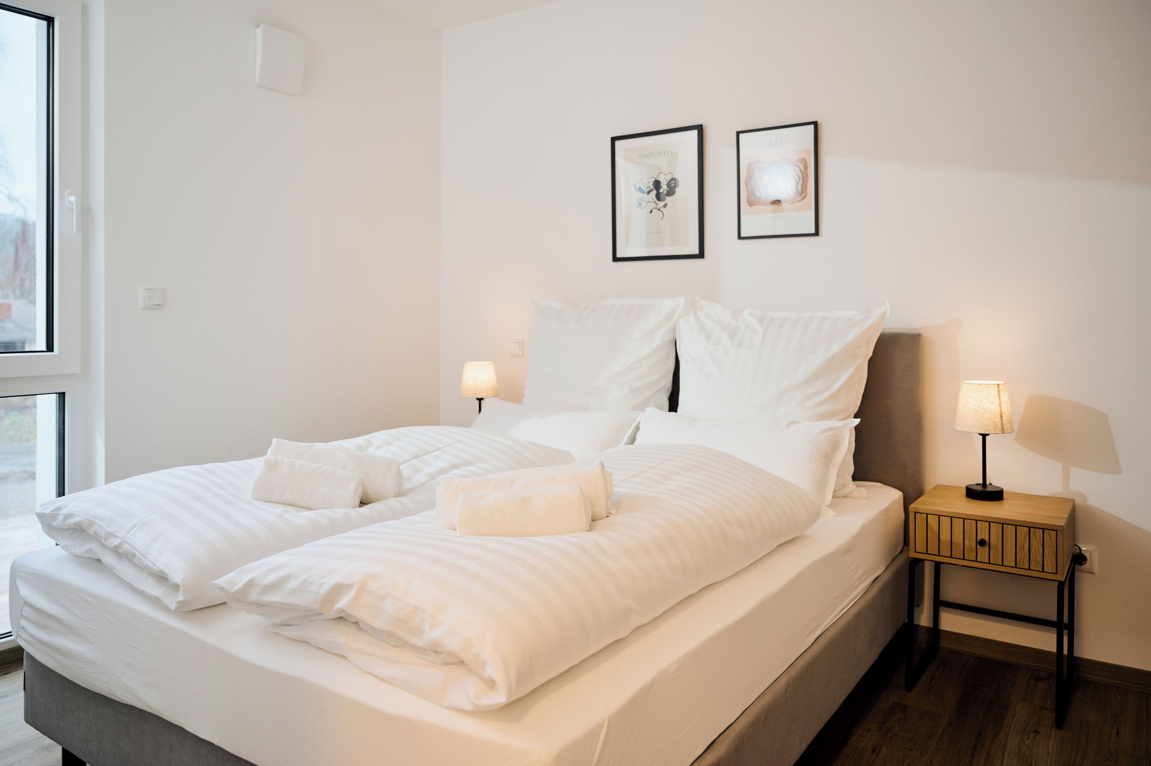 A neatly made bed with white linens, two bedside tables with lamps, and two framed pictures on the wall.