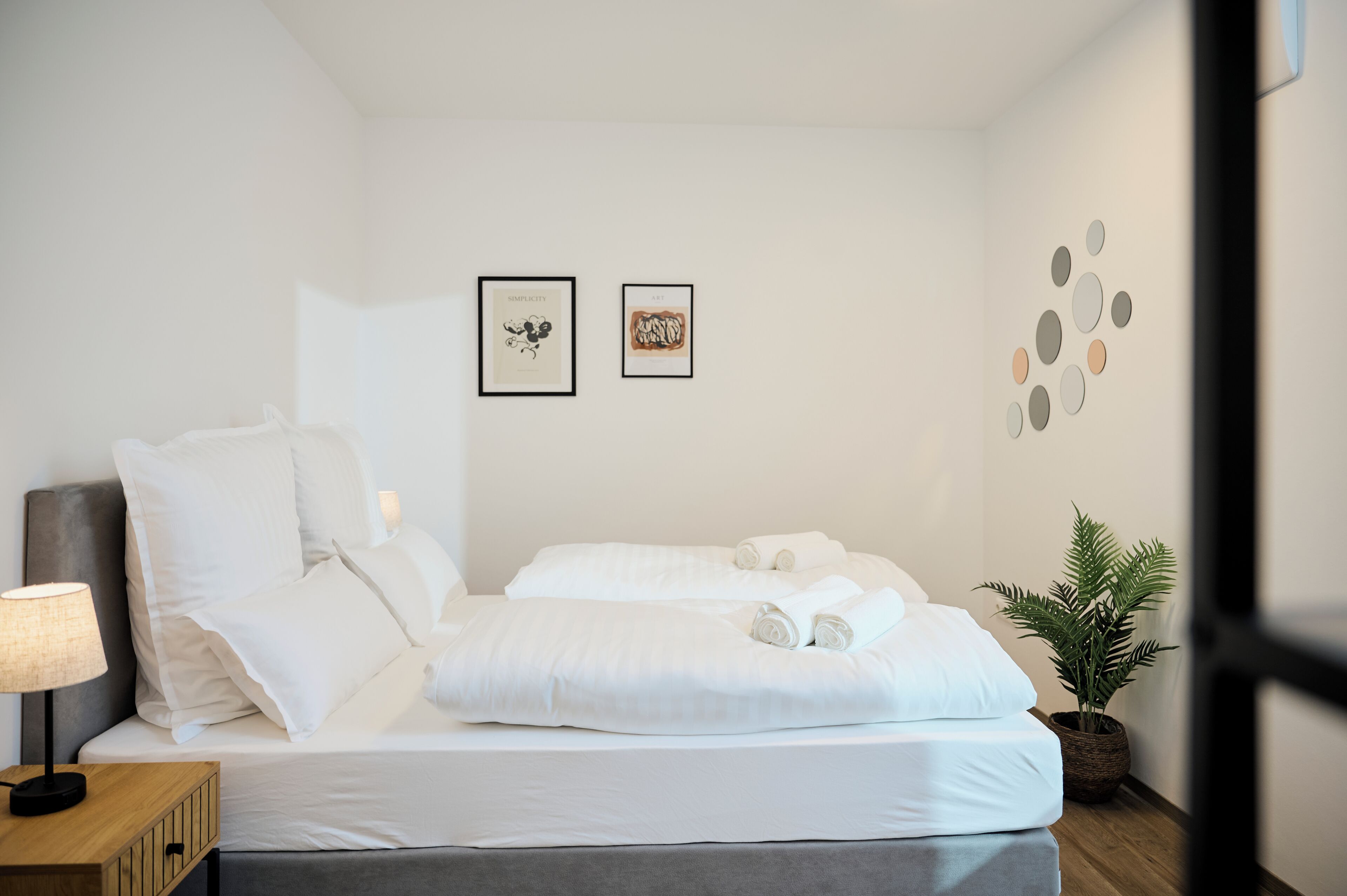 Ein ordentlich bezogenes Bett mit weißen Leinen, eine Nachttischlampe und ein gerahmtes Bild an der Wand.