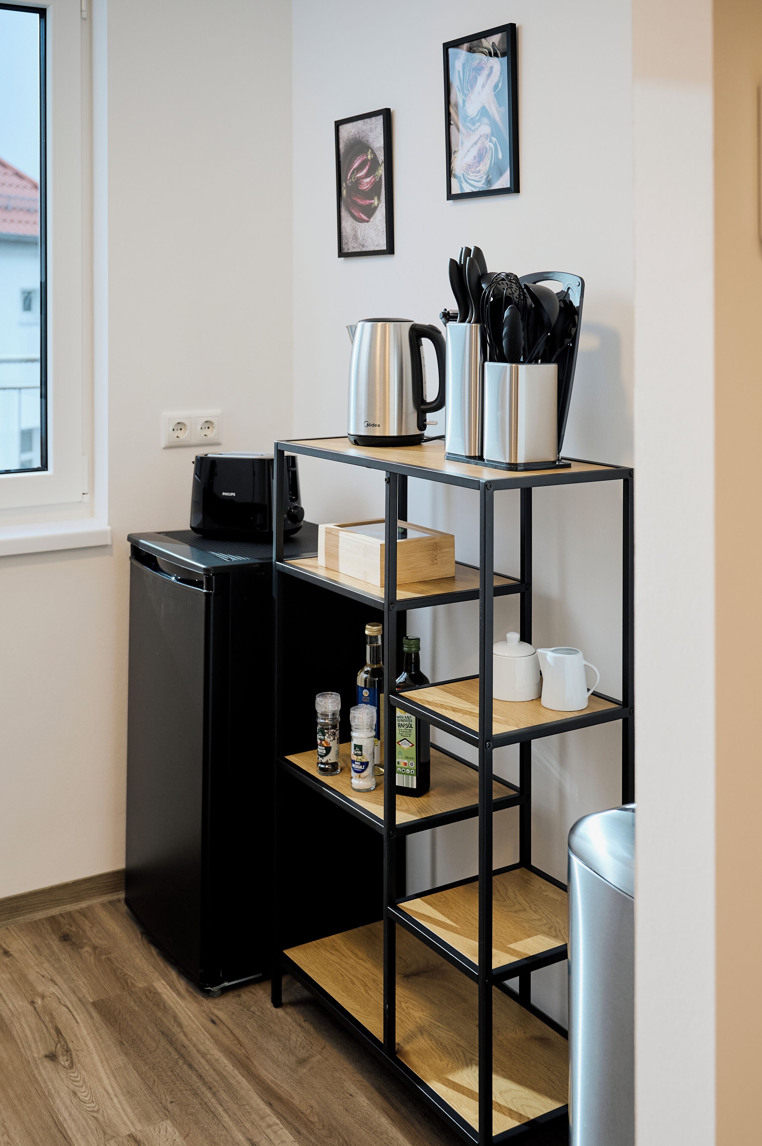 Une cuisine compacte équipée d’un réfrigérateur noir, d’une machine à café, d’une bouilloire et d’une étagère contenant des ustensiles de cuisine.