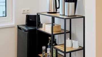 Une cuisine compacte équipée d’un réfrigérateur noir, d’une machine à café, d’une bouilloire et d’une étagère contenant des ustensiles de cuisine.