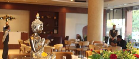A Buddha statue on a table in a restaurant setting with chairs and tables, a waiter in the background, and a large window with natural light.