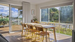 Uma sala de jantar moderna com uma mesa grande e cadeiras, janelas amplas com vista para vegetação e uma estrada, e uma porta de vidro deslizante para um espaço ao ar livre.