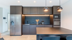A modern kitchen with a built-in refrigerator, oven, and microwave. There is a dining table with chairs and pendant lighting above. - Villa Memorias | Modern | 3 Bedrooms | Albufeira (Albufeira)