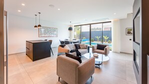 A modern living room with a sofa, coffee table, and dining area. There is a large window with a view of a swimming pool and greenery. - Villa Memorias | Modern | 3 Bedrooms | Albufeira (Albufeira)