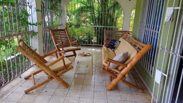 Un espace extérieur aménagé avec des sièges, abrité et doté de chaises en bois et d’une petite table, entouré d’une rambarde en métal et de verdure.