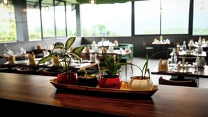 Een restaurantinterieur met gedekte tafels, plantenbakken op een dienblad en grote ramen met uitzicht op groen.