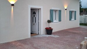 Ein Gebäude mit weißer Fassade, einer Tür und zwei Fensterläden. An beiden Seiten der Tür befinden sich jeweils zwei Wandleuchten.
