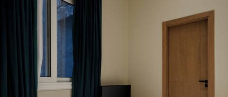 Un dormitorio con cama, televisor sobre una mesita de noche de madera, ventana con cortinas y una puerta de madera.