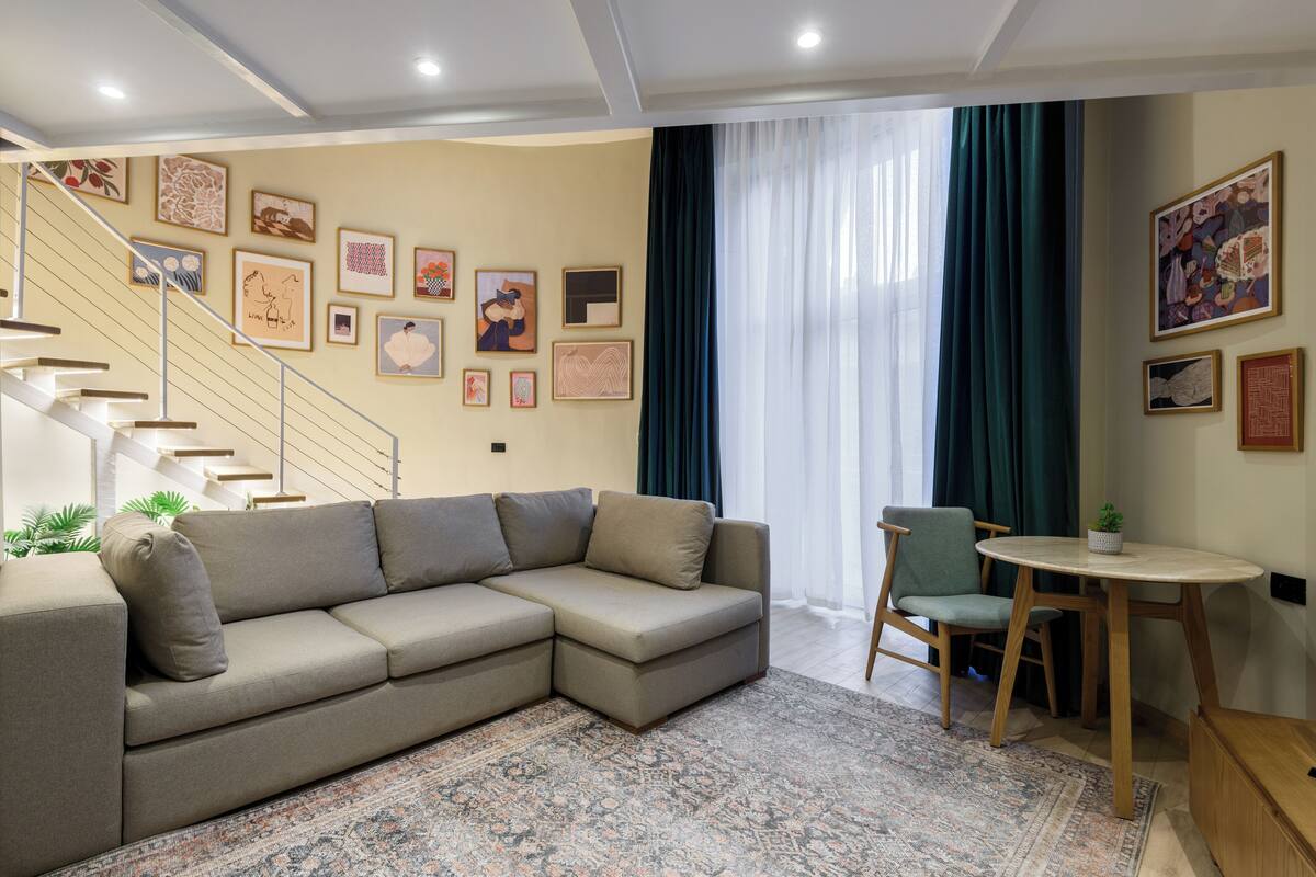 A living room with a sectional sofa, a dining table with chairs, a staircase, and artwork on the walls.