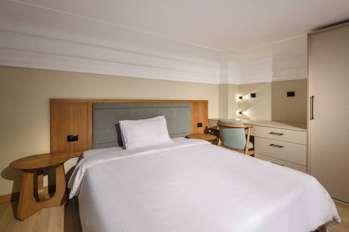 A hotel room with a bed, a wooden nightstand, a desk with a chair, a wardrobe, and a mirror with lights.