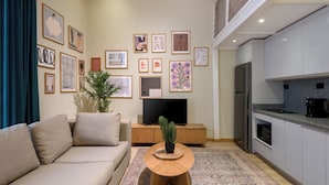 A modern living room with a sofa, coffee table, and a flat-screen TV. There are framed artworks on the wall and a kitchen area with a refrigerator and microwave. - Hazel Spaces DownTown (Cairo)